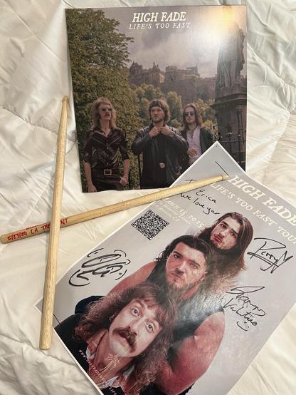 The image features a vinyl album cover titled "High Fade - Life's Too Fast" alongside a signed promotional poster of the band. Two drumsticks from the drummer are placed across the items, and the background is a white quilt. The poster includes a QR code and the band featured, along with autographs showing Erica love for her support. 
