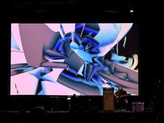pink grey blue and black shapes twisting on a screen, musician in front performing
