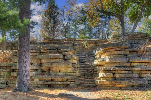Stone steps wind upward, nestled between large, layered rock formations surrounded by lush trees and fallen leaves.