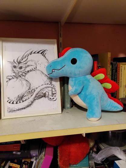 Ein cartoon-artiger blauer Plüsch-Dino mit rotem Rückensegel und Kopfschmuck in einem Bücherregal. Daneben in einem Bilderrahmen die Zeichnung eines Drachen. 