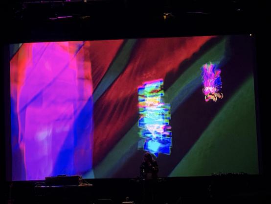 multicolored shapes overlayed and blurring on a movie screen, a musician on stage in front performing with a number of musical instruments
