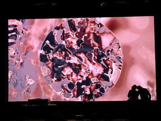 pinks greys and blacks shapes overlayed and blurring focused on a circle on a movie screen, two musicians on stage in front performing with a number of musical instruments on a table