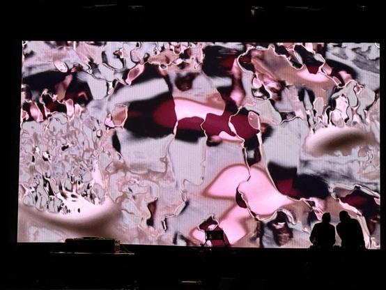 pinks greys and blacks shapes overlayed and blurring focused on a circle on a movie screen, two musicians on stage in front performing with a number of musical instruments on a table