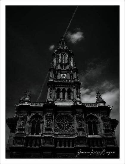 
The image presents the architectural structure of the Holy Trinity Church in Paris, standing tall and imposing against a dramatic sky. The building, reminiscent of a historical or religious edifice, features intricate details and ornate carvings that hint at its grandeur and significance. Its dark, monochromatic palette evokes a sense of mystery and solemnity, enhancing the intricate textures of the facade.
The towering spire, adorned with statues, draws the eye upward, leading to an almost celestial backdrop where wisps of clouds and a jet trail create lines that add depth and movement to the scene. The play of light and shadow accentuates the contours of the building, highlighting its architectural features and giving it a timeless quality.