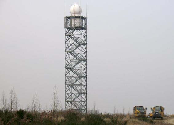 Ein Turm aus Stahl, oben ein weißer Container, darüber ein kugelförmiger Radom, umgeben von vier Blitzableitern. Rechts unten am Bildrand zwei LKW.