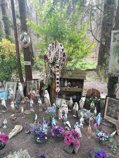 Photo d'un parterre de fleurs, remplis de statuettes de vierge Marie, une grande croix en bois recouverte de chapelets