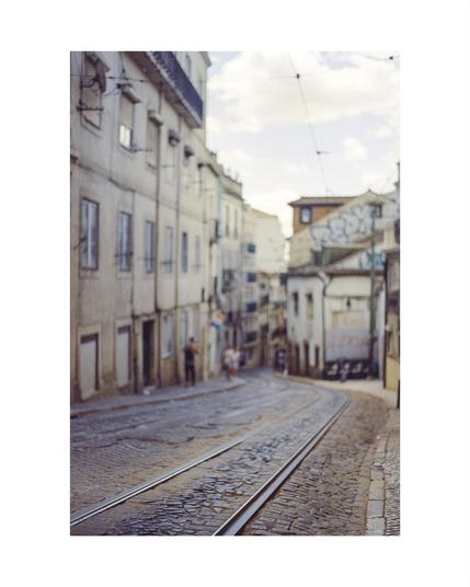 empty tram tracks in the middle of a paved road in lisbon, in blurry background buildings