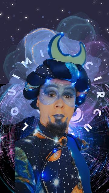A close-up portrait of a performer in a cosmic costume, featuring a headpiece with a glowing crescent moon. Their blue-painted face, adorned with starry makeup, is set against a futuristic space-themed background with floating lights and digital textures.