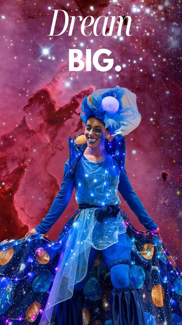 A stilt-walker dressed in a luminous blue outfit spreads an expansive cape decorated with planets and LED lights. Their radiant smile, cosmic makeup,