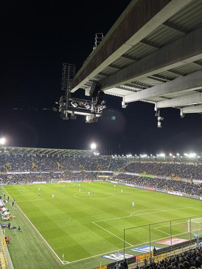 A nighttime image of a football match in a stadium. It's Club Brugge against Charleroi. The field is illuminated, with players in action and a large crowd visible in the stands. Equipment, including cameras and lights, is mounted from the stadium roof, highlighting the vibrant atmosphere of the game