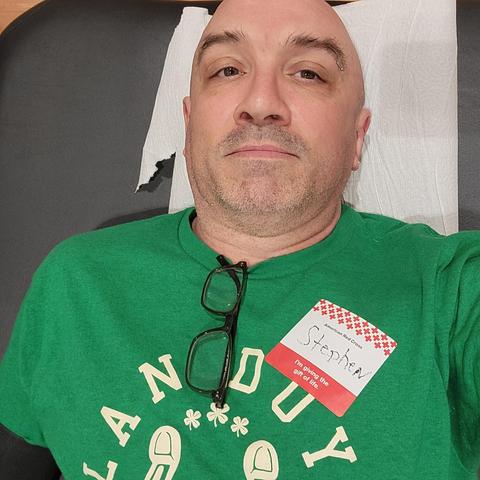Picture of me in a green shirt. With a red and white name tag laying down on a bed face up donating blood.