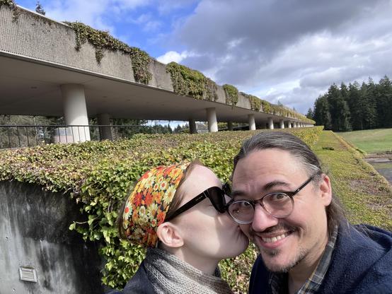 Photo of me, a 40-something white lady, kissing my husband, a 40-something white guy, on the cheek. He looks alarmed, maybe? But not displeased. (I think.) Behind us, the old Weyerhaeuser campus. 
