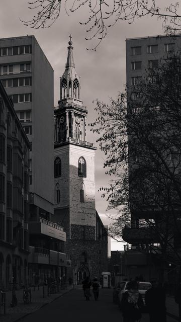 A black and white photograph of St. Mary's Church in Berlin, framed by modern buildings. The sun breaks through the clouds, casting light on the historic church tower. Leafless tree branches and people walking on the street add depth to the scene.