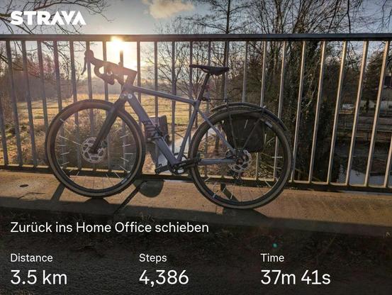 Bild aus einer Fitnessapp. Es zeigt ein Gravelbike, das an einem Metallgeländer lehnt, die Sonne scheint durch die Streben des Geländers. Der Text ist "Zurück ins Home Office schieben"