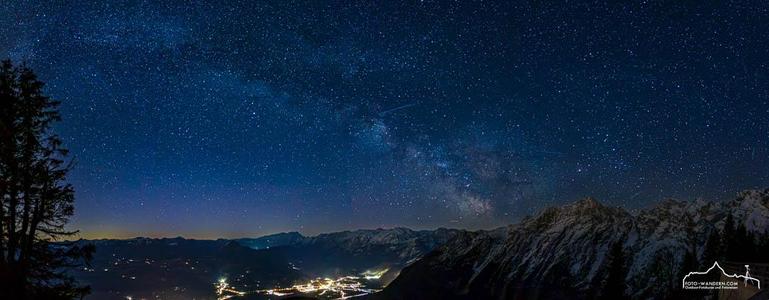 Das Bild zeigt eine Nachtansicht einer Stadt zwischen Bergen, im Salzburger Land wobei der Himmel mit Sternen bedeckt ist und die Milchstraße von rechts über die Berge durch das Bild führt