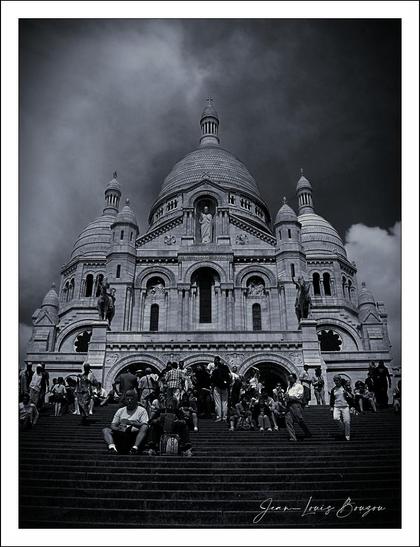 In this black and white image, the intricately designed facade of the Sacré-Coeur baislica rises dramatically against a moody sky. The domes and spires of the building create a majestic silhouette, evoking a sense of timelessness and reverence.
The stairs leading up to the church are bustling with people, each person a small part of the larger story unfolding around this iconic landmark. Their presence adds a sense of life and vibrancy, contrasting with the stillness of the architectural masterpiece above.
The clouds hover overhead, hinting at a potentially dramatic change in weather, which could symbolize the contrast between light and darkness—both literally and metaphorically. The image captures not only a place of worship but also a gathering point for diverse individuals, suggesting themes of community and exploration.
This composition bears a certain timelessness, inviting the viewer to ponder the narratives of those who visit, as well as the layers of history embedded within the walls of the church itself. The interplay of architecture, the natural environment, and human activity creates a rich tableau, encouraging reflection on both the sacred and the everyday.