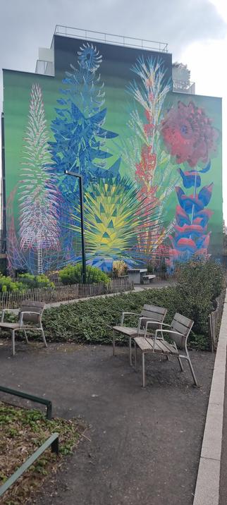 Une fresque murale colorée représentant des plantes stylisées aux formes géométriques et aux teintes vives (bleu, rouge, jaune, vert) recouvre la façade d’un immeuble. Devant le mur, un petit jardin avec des bancs et des chaises en bois est aménagé, entouré de végétation.