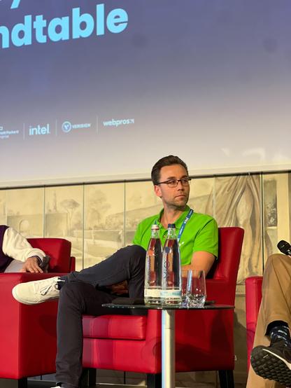 Ronny Marx, Commercial Director at Syde, sits on a red chair on stage during a panel discussion. He is wearing a green polo shirt and a blue lanyard. In front of him, two water bottles are placed on a small table. A large screen in the background displays event sponsors.