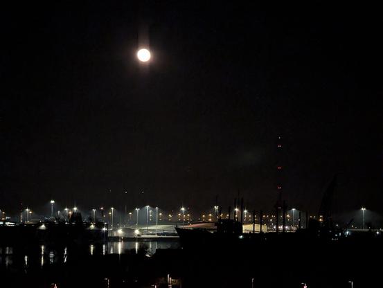Es ist dunkel. Der Hafen ist nur an der Beleuchtung zu erkennen, die sich teilweise im Fluss spiegelt. Das Wasser selbst ist tiefschwarz ebenso wie der Himmel. Über dem Hafen steht der Mond fast voll.