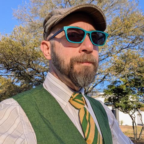 A photo of me, a 47 year old white man with a shaved head, a short cropped brown & grey beard, 

photographed from the chest up

In front of some live oak trees in an office park,

wearing

rectangular green acetate glasses, a green and gold diagonally striped tie in a trinity knot  on  brown pinstriped dress shirt under a green vest with a tan flat cap.