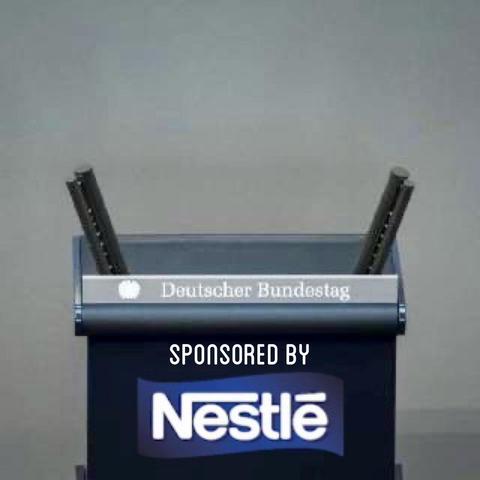 Ein Podium mit dem Text "Deutscher Bundestag" darauf, auf dem zwei Mikrofone befestigt sind. Das Bild enthält den Satz "Gesponsert von Nestlé", der prominent unten angezeigt wird. Der Hintergrund ist schlicht und neutral.