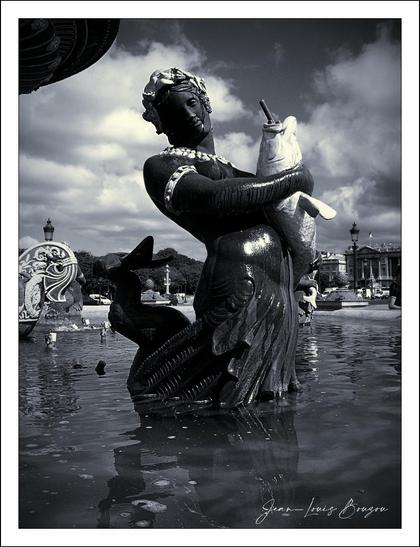 The image showcases a striking sculpture of a mermaid-like figure standing in the Fountain of the Seas at Concorde Square in Paris. The genie is gracefully poised, embracing a fish with a gentle yet confident demeanor. Her hair is adorned, adding to the ethereal quality of the piece, while the intricate details of her tail suggest both elegance and strength.
Surrounding him is a tranquil aquatic environment, with reflections on the water's surface enhancing the overall mystique. The black and white treatment of the image adds a timeless quality, emphasizing the sculpture's textures and forms without the distraction of color.