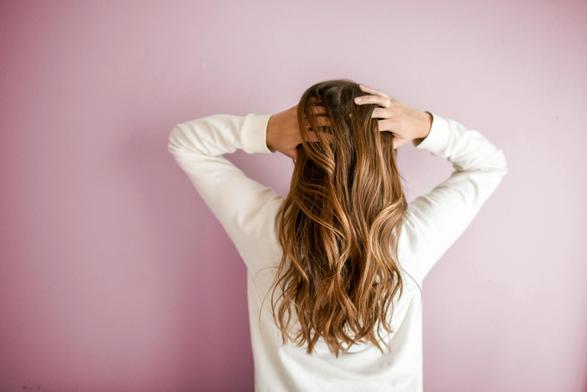 A person with long, wavy brown hair stands with their back to the camera, lifting their hair with both hands. They are dressed in a white, long-sleeve shirt, positioned against a soft, light pink wall.
