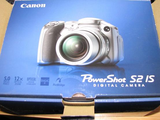A cropped photo of the vintage digital camera's box taken with the vintage camera with flash on. It shows it is a Canon PowerShot S2 IS Digital Camera with some features highlighted and an image or rendering of the camera. The box is mostly blue and the camera is silver and gray.