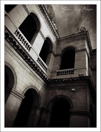 This image captures a striking architectural perspective, with an emphasis on the upward gaze towards the intricately designed arches and balustrades. The monochromatic tones create a dramatic atmosphere, enhancing the interplay of light and shadow. The stonework, with its textured surfaces, speaks to a sense of history and permanence.
The angles guide the viewer's eye, inviting exploration of the depth and dimensions among the columns and arches. There’s a contrast between the solidity of the structure and the somewhat ominous clouds overhead, suggesting themes of grandeur entwined with mystery or unease. This juxtaposition can evoke feelings of introspection, prompting thoughts about the passage of time and the stories embedded within the stone.
