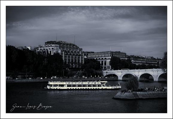 In this evocative image, a tranquil scene unfolds along the banks of a river, the Seine, given the context. The black and white palette adds a timeless quality, accentuating the stark contrasts between light and shadow.
A gently cruising boat, illuminated by warm lights, stands out against the cooler tones of the landscape, suggesting a sense of life and movement amid the stillness. The reflections on the water create a shimmering texture that draws the eye, inviting one to linger on the scene.
In the background, stately buildings rise gracefully, their architectural details softened by the monochromatic scheme. The presence of trees adds a touch of nature, balancing the man-made structures and hinting at a connection between urban life and the natural world.

