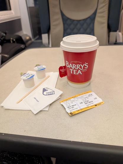 A table in a train with two creamers, two packets of sugar, a napkin, stirrer, a cup of Barry's Tea, and a train ticket from Cork, Ireland to Dublin, Ireland.