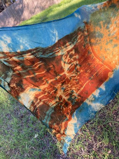 A large rectangular shawl blowing in the wind. The edges are blue and the center is rust with an impression of a dandelion. 