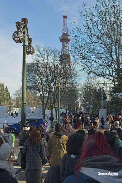 a photograph taken in Sapporo, at the Snow Festival, 2025.  The photo looks over a crowd as they walk towards the Sapporo tower in the distance.  You can see a few snow sculptures on the left.