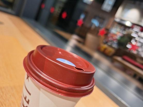 Close up shot of the top of a take away coffee cup, standing on a counter in front of a dark window. In the window there is a blurry reflection of the coffee shop interior (some tables, a cooling shelf)