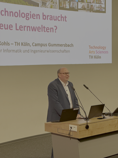 Professor Kohls von der Technischen Hochschule Köln am Pult des Hörsaals 1. Im Hintergrund sieht man die Projektion einer Folienpräsentation auf dem zentral der Begriff Lernwelten gefolgt von einem Fragezeichen zu lesen ist.