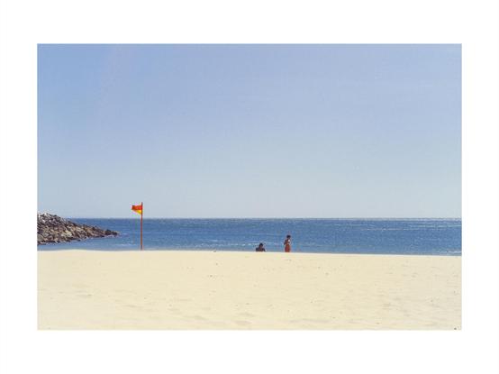 view of a beach with yellow sand, blue water and sky, blurry line between sand and blue, warning flag blowing in the wind