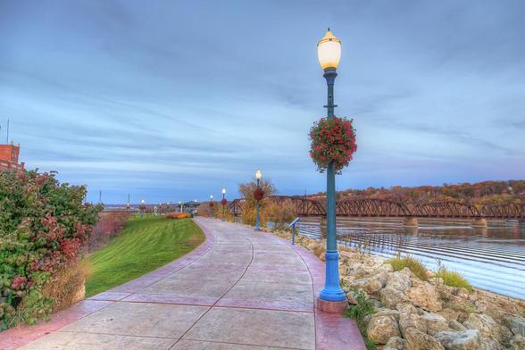 A peaceful riverside path is lined with lampposts adorned with hanging flower baskets, creating a scenic walkway. The calm water reflects the blue hue