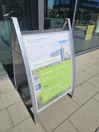 Ein Aufsteller vor dem Eingang des ITMC in Dortmund mit einem Poster, auf dem "Herzlich Willkommen" steht, und zu den beiden Tagungen ZKI AK Web und Confluence.campus eingeladen wird.