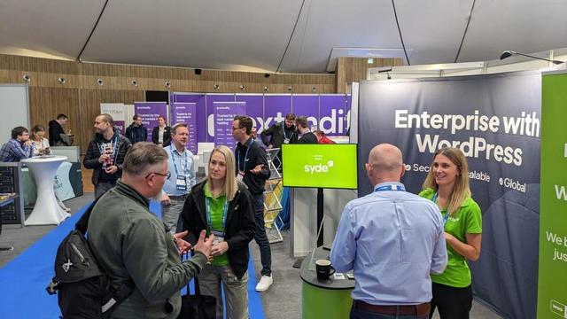 Various visitors and Syde employees in green shirts are standing at the Syde booth at CloudFest, discussing WordPress solutions. Other exhibition stands are visible in the background.