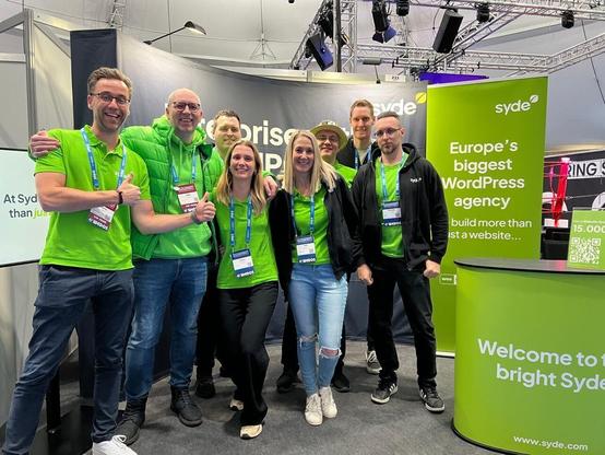A group of Syde employees in green shirts poses smiling for a group photo at their booth at CloudFest. Exhibition banners and lighting can be seen in the background.
