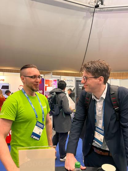 A Syde employee in a green shirt is talking to a visitor in a suit at the Syde booth at CloudFest. Both are making friendly eye contact and having an engaging discussion.
