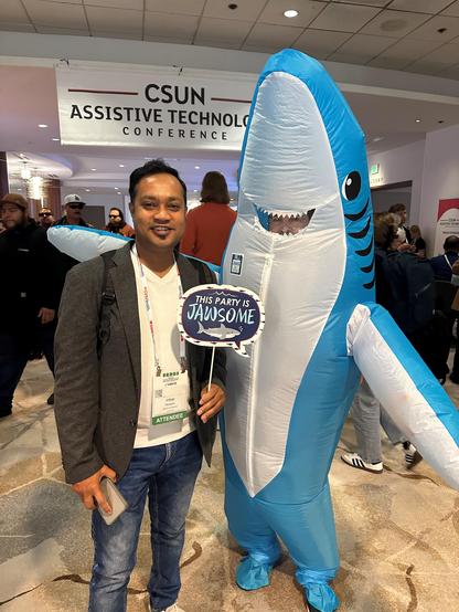 Imtiaz standing with Sharky in front of the CSUN Assistive Technology Conference banner.