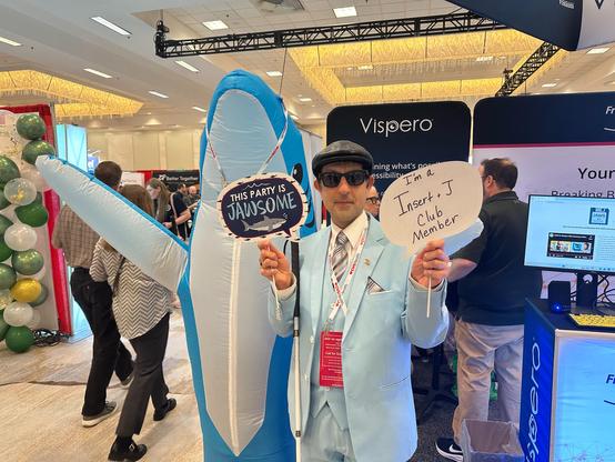 Man standing with Sharky in the exhibit hall while holding two signs, one reads "This party is Jawsome!" and the other reads "I am an Insert J Club member."