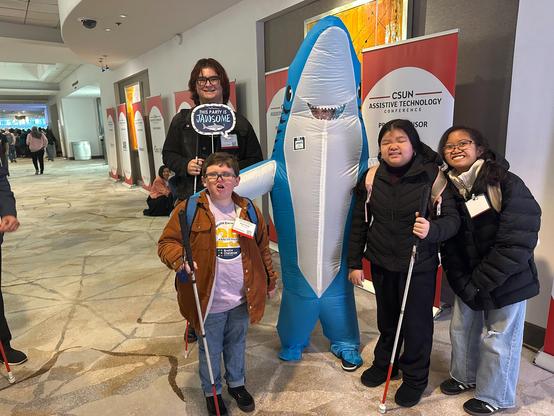 A group of four young people standing with Sharky in front of the exhibit hall.