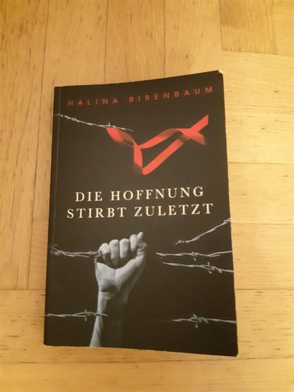 Bild eines Buchcovers: Eine nach oben gestreckte Hand vor schwarzem Hintergrund. Darin ein Stacheldraht. Darüber ein rotes Band. - Text oben: "Halina Birenbaum" Text in der Mitte: "Die Hoffnung stirbt zuletzt"