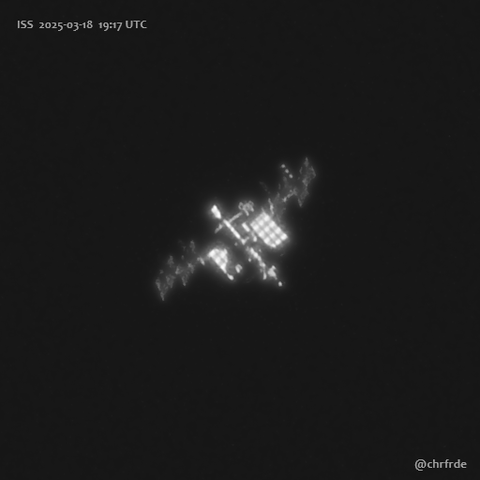 Telescopic view of ISS against black background. Direction of movement in this image appears at an angle 45 degree towards the top left. Solar panels are dimly visible but foreshortened being seen at a steep angle. Crew-10 Dragon capsule docked at the forward port appears as a bright elongated object borderline resolved to show a bit of tapering at the nose.