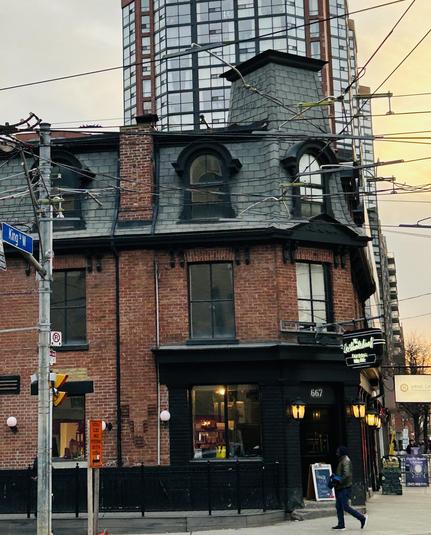 A historic brick building with a sloped roof and large windows is situated at the corner of a street. Modern glass skyscrapers loom in the background, and electrical wires run overhead. A person walks past the entrance of the building, which features a sign.