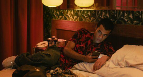 Image tirée de “Baby”, de Marcelo Caetano : dans une chambre aux rideaux tirés, le personnage principal, un jeune homme, est allongé sur le lit, son smartphone à la main.