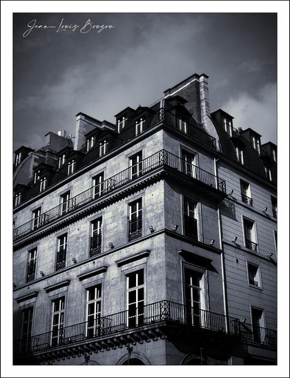 The image presents a building with a distinctive architectural charm, captured in a monochromatic palette. The façade exhibits elegant symmetry, with numerous windows framed by delicate iron railings. The building rises sharply, its upper levels accentuated by steep, sloping roofs that add a dramatic touch to its silhouette.
Clouds swirl in the background, creating a moody atmosphere that contrasts with the solid, timeless structure. This interplay between light and dark evokes a sense of nostalgia, perhaps suggesting stories of the past embedded within the walls.
The choice of black and white emphasizes texture and form, drawing attention to the intricate details of the building's design. The artist's signature is subtly included, hinting at a personal connection or interpretation of this scene. The overall effect speaks to themes of history, permanence, and the architectural beauty found in urban landscapes.