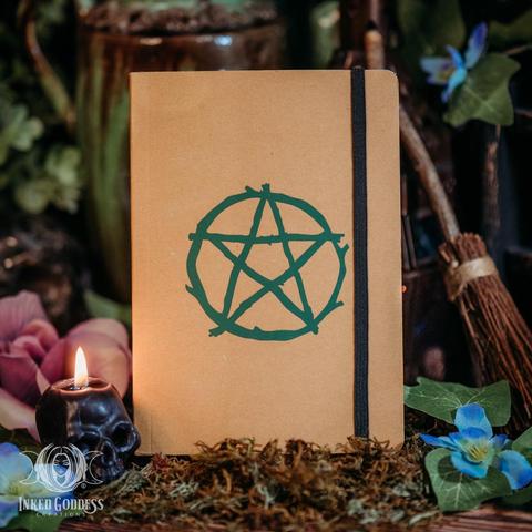 A small kraft paper-brown "Pentacle Branch Eco-Friendly Notebook" is shown with its cover closed. On the cover, a pentacle that looks as if it was made out of branches in the center in a solid forest-green silhouette. There is a black elastic band lengthwise on the right side to hold the notebook closed or bookmark to a page. This is an exclusive product from Inked Goddess Creations.
